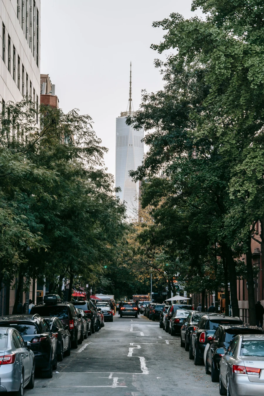 Tree-lined New York City street with parked cars and One World Trade Center in the background